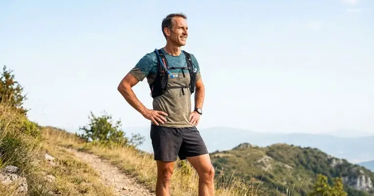 A smiling man stands confidently on a grassy mountain trail, wearing a gray shirt, black shorts, and a hydration pack. The background features hazy hills under a clear blue sky.