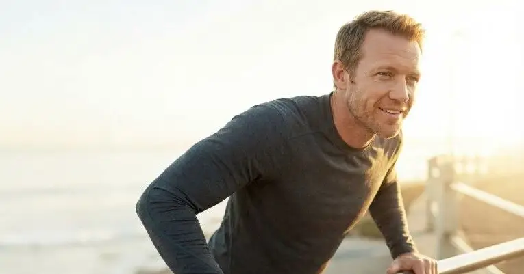 A man in a long-sleeve shirt leans against a railing by the beach, with the sun setting in the background.