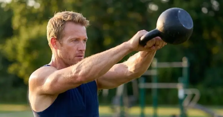 A person in a sleeveless shirt performs a kettlebell swing in an outdoor fitness area, surrounded by trees and gym equipment.